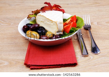 aged camembert cheese on green salad in white dish over cloth with olives and over wooden table