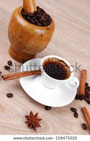 sweet hot drink : black arabic coffee in small white cup with mortar and pestle , beans spilled over wooden table , decorated with cinnamon sticks and anise stars