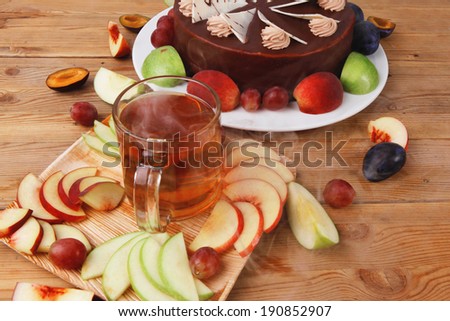 big whole frost chocolate cream brownie cake topped with white chocolate and cream flowers with hot tea cup decorated with fruits apple plum and grape on plate on wooden table