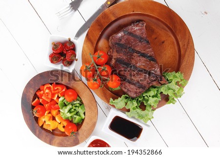 dinner of fresh rich juicy grilled beef meat steak fillet with marks on wooden plate over white table served with vegetable salad and cutlery, new york styled cuisine
