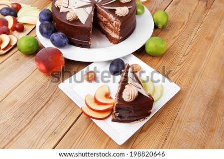 chocolate cream brownie cake layers slice topped with white chocolate and cream flowers decorated with fruits apple plum and grape on plate on wooden table with big whole cake
