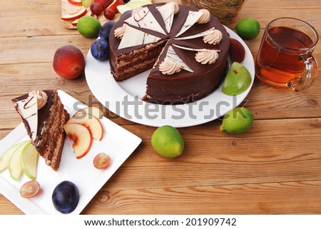 chocolate cream brownie cake layers slice topped with white chocolate and cream flowers decorated with fruits apple plum and grape on plate on wooden table with big whole cake