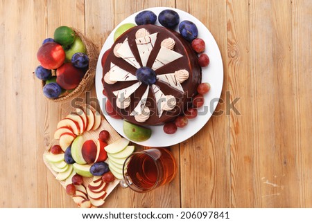 whole big chocolate cream brownie cake topped with white chocolate and cream flowers with hot tea cup decorated with fruits apple plum and grape on plate on wooden table