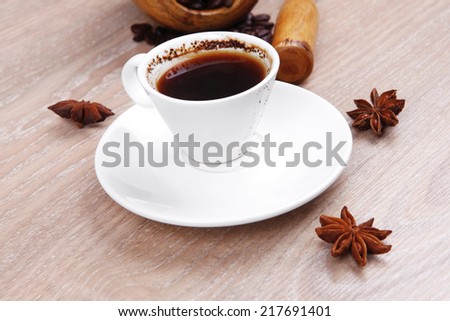 sweet hot drink : black arabic coffee in small white cup with mortar and pestle , beans spilled over wooden table , decorated with cinnamon sticks and anise stars