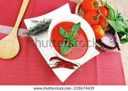 cold fresh diet tomato soup with basil thyme and dry pepper in big bowl over red mat on wood table ready to eat