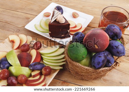 slice of chocolate cream brownie cake topped with white chocolate and cream flowers with hot tea mug decorated with fruits apple plum and grape on plate on wooden table