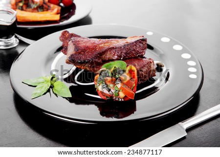 fresh red beef meat steak barbecue garnished vegetable salad sweet potato and basil on black plate over black wooden table with bbq sauce in sauceboat