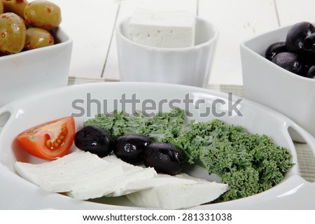 fresh greek feta cheese sandwich on white plate with hot green olives and black, goat cheese cube, tomatoes kale over retro table