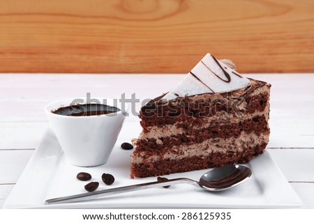 breakfast hot coffee mug and cream chocolate layer cake decorated with white chocolate slice and cream flower on white plate over wood