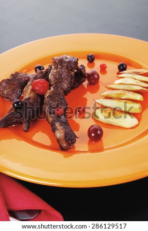 grilled beef meat with berries fried potatoes and cherry under sweet honey sauce on orange plate over black wooden table