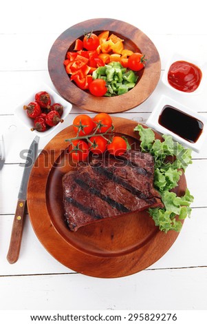dinner of fresh rich juicy grilled beef meat steak fillet with marks on wooden plate over white table served with vegetable salad and cutlery, new york styled cuisine