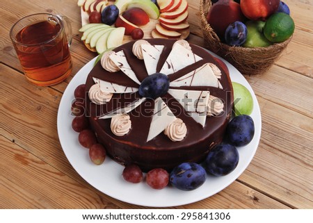 whole big chocolate cream brownie cake topped with white chocolate and cream flowers with hot tea cup decorated with fruits apple plum and grape on plate on wooden table