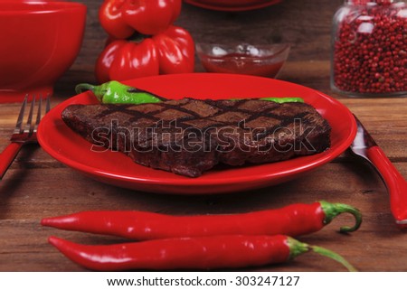 red theme lunch : fresh grilled bbq roast beef steak red plate green chili tomato soup ketchup sauce paprika small jug glass ground pepper american peppercorn and modern cutlery served on wooden table