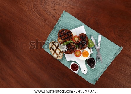 junk food meat big beef hamburger fried eggs on white  wood plate with cutlery ketchup sauce and pickles on blue mat over wooden table