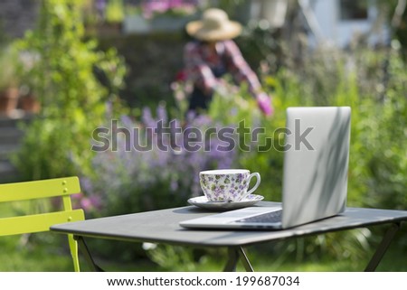 Woman in a garden, focus on cup of tea and laptop foreground