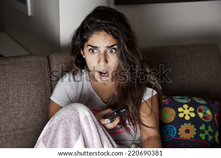 Scared young woman watching tv. Brunette girl sitting on couch with remote control