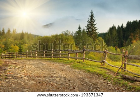 Autumn landscape in a mountain valley: road and wooden fence at sunlight