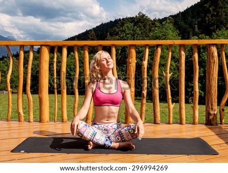 Woman sitting in lotus pose doing yoga over mountains, green field and blue sky background