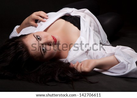 Young woman with long dark hair and red lips, in the white men's shirt with a sensual look lies on his back on the ground, on a black background