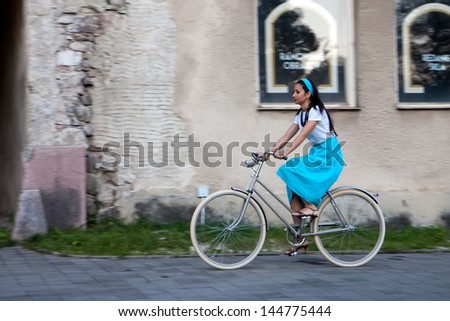 A woman in a turquoise skirt with a scarf on her head rides a retro bike next to old building