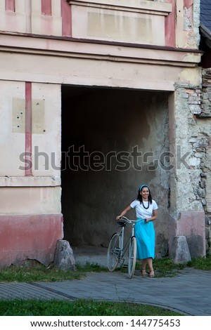 A woman in a turquoise skirt with a scarf on her head comes next to retro bicycle from old building