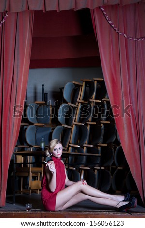 Actress young lady in red dress sits sad with a cigarette and a glass of alcohol on a dusty old stage