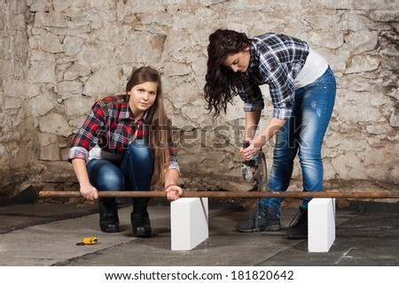 Two young long-haired woman cut iron pipe with angle grinder during the repair an old house