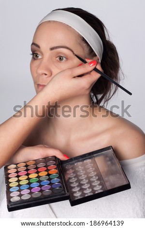 Beautician holding in one hand palette cosmetic shadows, and using a brush to apply color to the eyebrows of young brunette woman with headband on her head