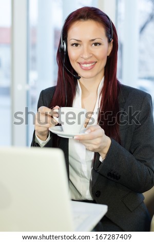Woman with headphone talking,using a laptop computer and holding a cup of hot drink