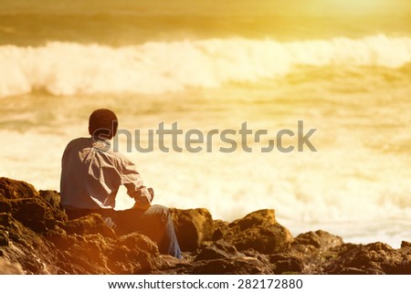 Rear  view of pensive, depressed man sitting on coast rock, looking at sea horizon.Copy space, unrecognizable person, lens flare, sunset light