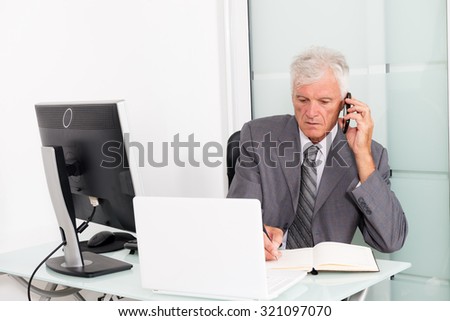Senior businessman using a cell phone and writing an information on a document in his office