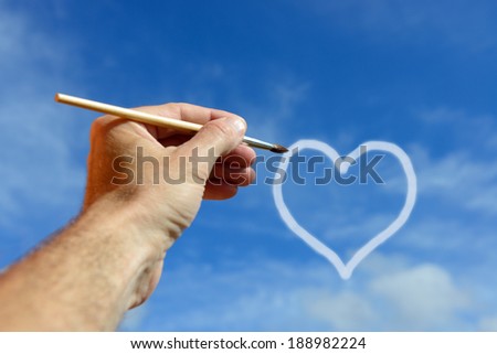 Hand with brush draws heart on the blue sky
