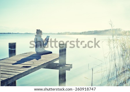 Washed out photo look. Girl reading from a tablet on the wooden jetty against a lake. Switzerland