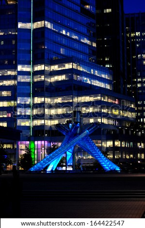 The Vancouver Canada Olympic Torch and Cauldron on display at night against tall skyscrapers.