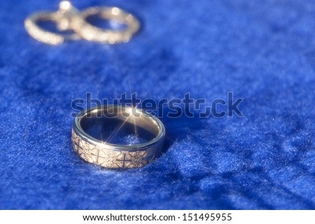 Groom\'s wedding band with bride\'s wedding and engagement rings in the background, laying on bright blue fabric.
