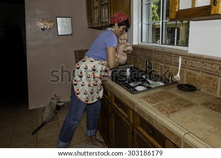 A retro-style homemaker and mother removes happy baby from kitchen sink after giving baby a bath while cat observes