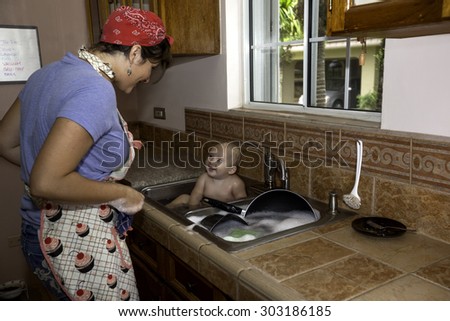 A retro-style homemaker and mother gives happy baby a bath while doing the dishing in kitchen double sink