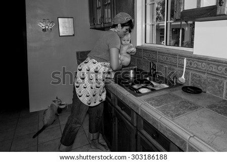 A retro-style homemaker and mother removes happy baby from kitchen sink after giving baby a bath while cat observes; in black and white