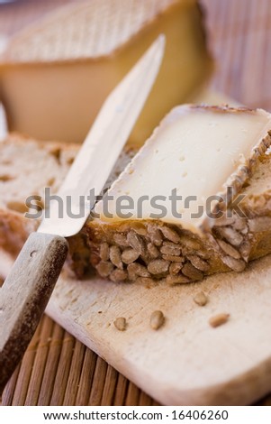 A french tasty slice of cheese on a piece of bread with cereals on a wooden board, with the whole cheese and a knife in the foreground