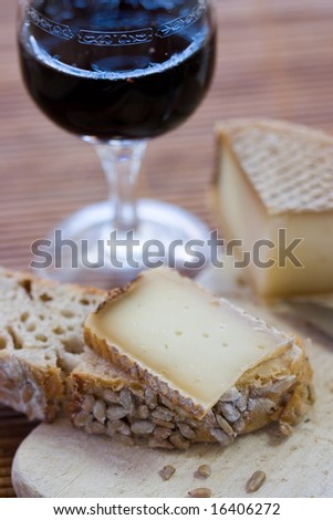 A french tasty slice of cheese on a piece of bread with cereals on a wooden board, with the whole cheese and a glass of red wine and a knife in the background