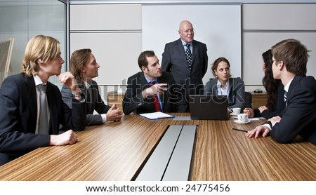 An office meeting between a senior executive and six of his junior staff members