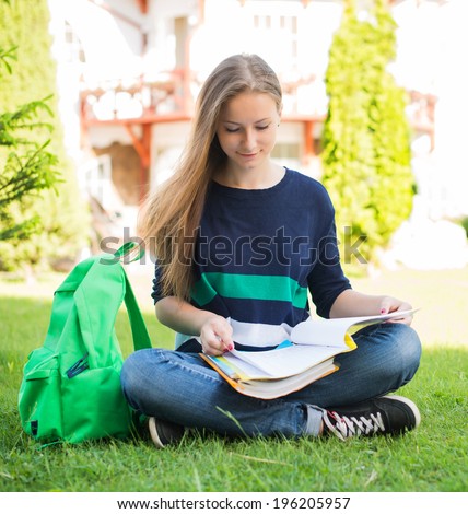 Beautiful school or college girl sitting on the grass with books and bag studying in a park.