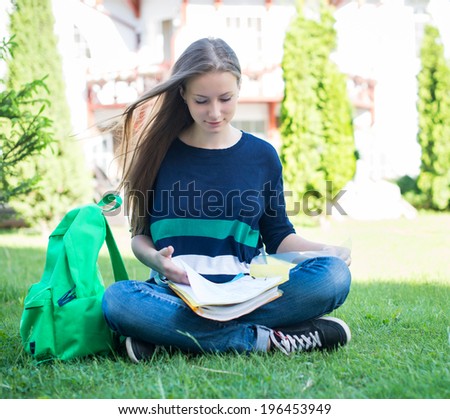 Beautiful school or college girl sitting on the grass with books and bag studying in a park.