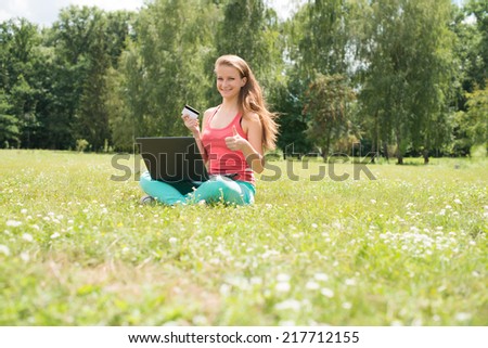 Internet shopping woman online with laptop and credit card sitting outdoor on green grass. Happy internet shopper with thumb up buying things on the internet.