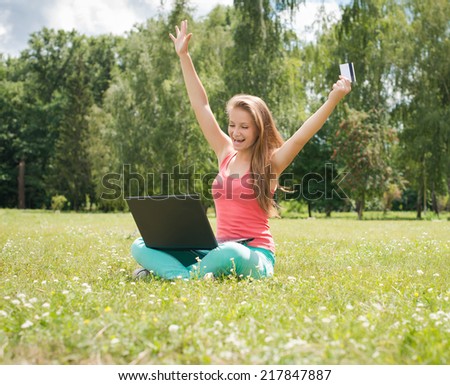 Happy internet shopping woman online with laptop and credit card sitting outdoor on green grass. Internet shopper buying things on the internet.
