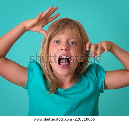 Angry little girl growls isolated on turquoise background. Portrait of child making a face and grimacing at camera.