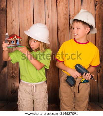 Little builders in helmet with a house model. Family house concept - portrait of lovely boy and girl with house and tools on wooden background. Concept of real estate and engineering.