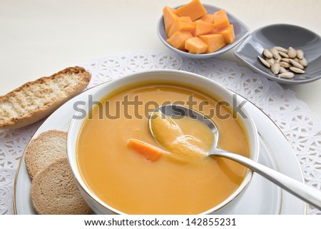 Pumpkin squash soup and spoon, all exhibited in table with tablecloth.