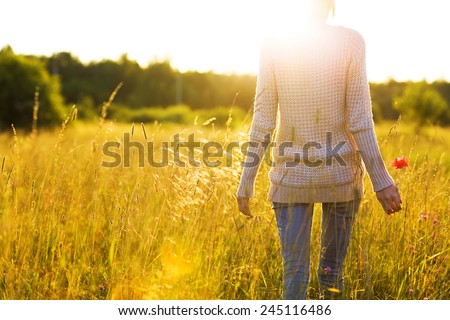 Young woman walking in the field toward the sun holding a poppy flower.