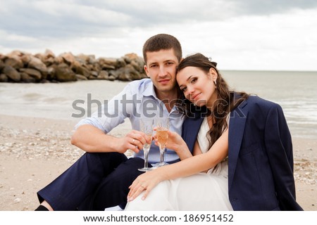 Happy newlyweds sitting on the beach with glasses of champagne. Loving bride and groom near the sea.
